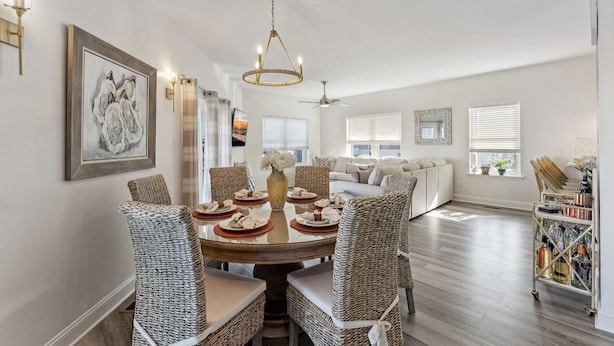 A dining area with a round table, wicker chairs, and a chandelier. There is a sofa and a window in the background.