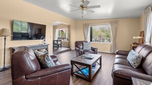 A living room with leather sofas, a coffee table, a ceiling fan, and a TV mounted on the wall. - NW Tucson family friendly house with Catalina mountain views (Tucson)