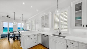 A modern kitchen with white cabinetry, a dishwasher, and a large island. There is a dining area with a table and chairs, and a view of the ocean through large windows. - BellaVista on the Beach*HotTub*Gulf Front*3 Patios (Miramar Beach)