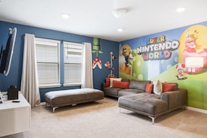 A room with a Nintendo-themed mural, a grey sofa, a white desk, and a window with blinds.