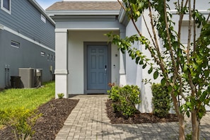 A residential building with a grey door, a small front yard with a brick pathway, and a tree.