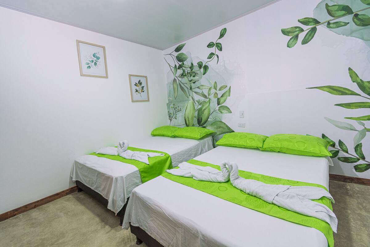 A hotel room with two beds, each with green pillows and white linens. There are framed botanical prints on the walls.