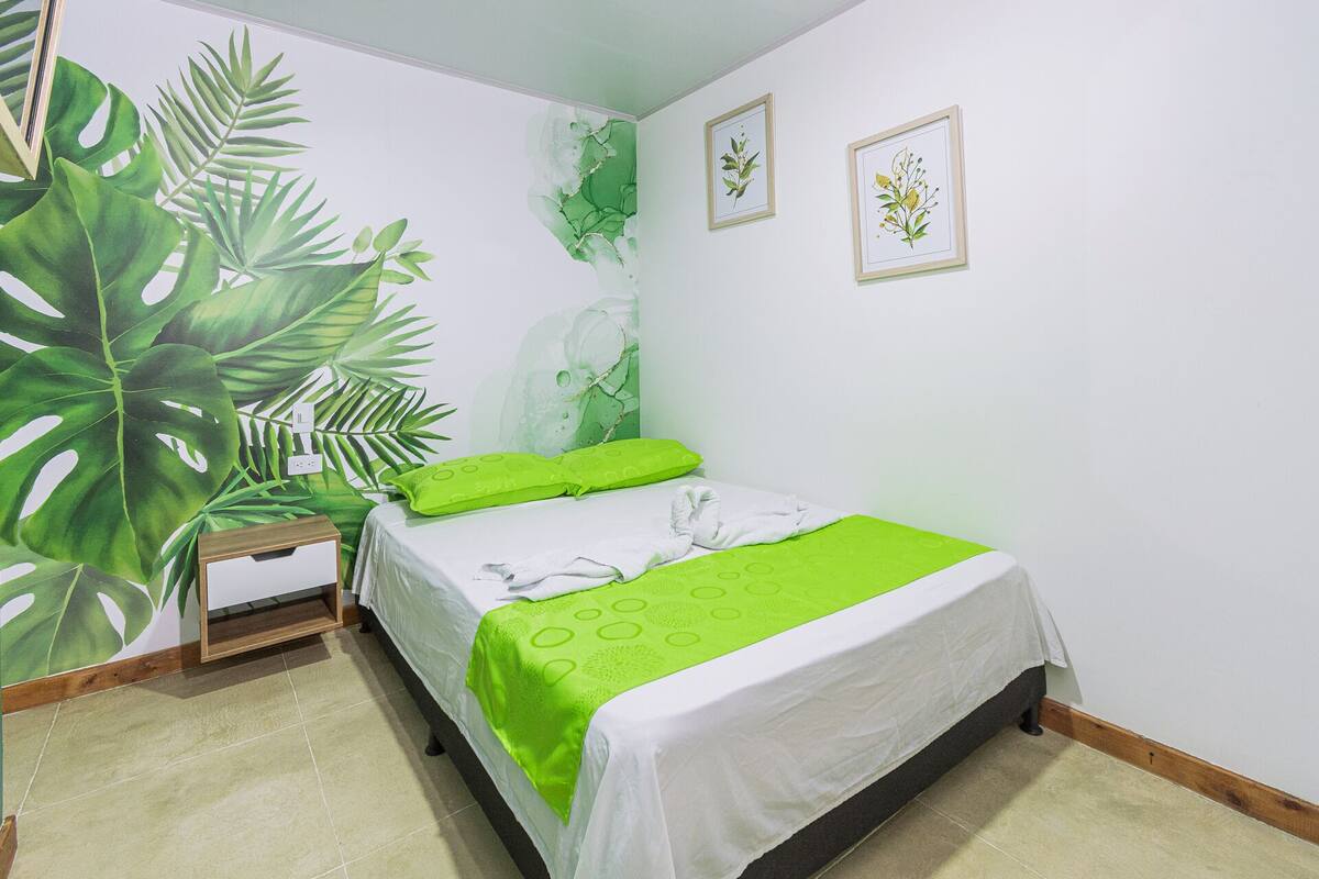 A bedroom with a bed, green pillows, a nightstand, a wall with a plant mural, and two framed botanical illustrations.