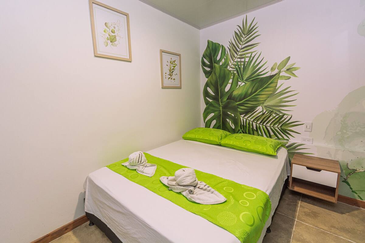 A neatly made bed with green bedding and a towel, a nightstand with a lamp, and a large plant in the corner.