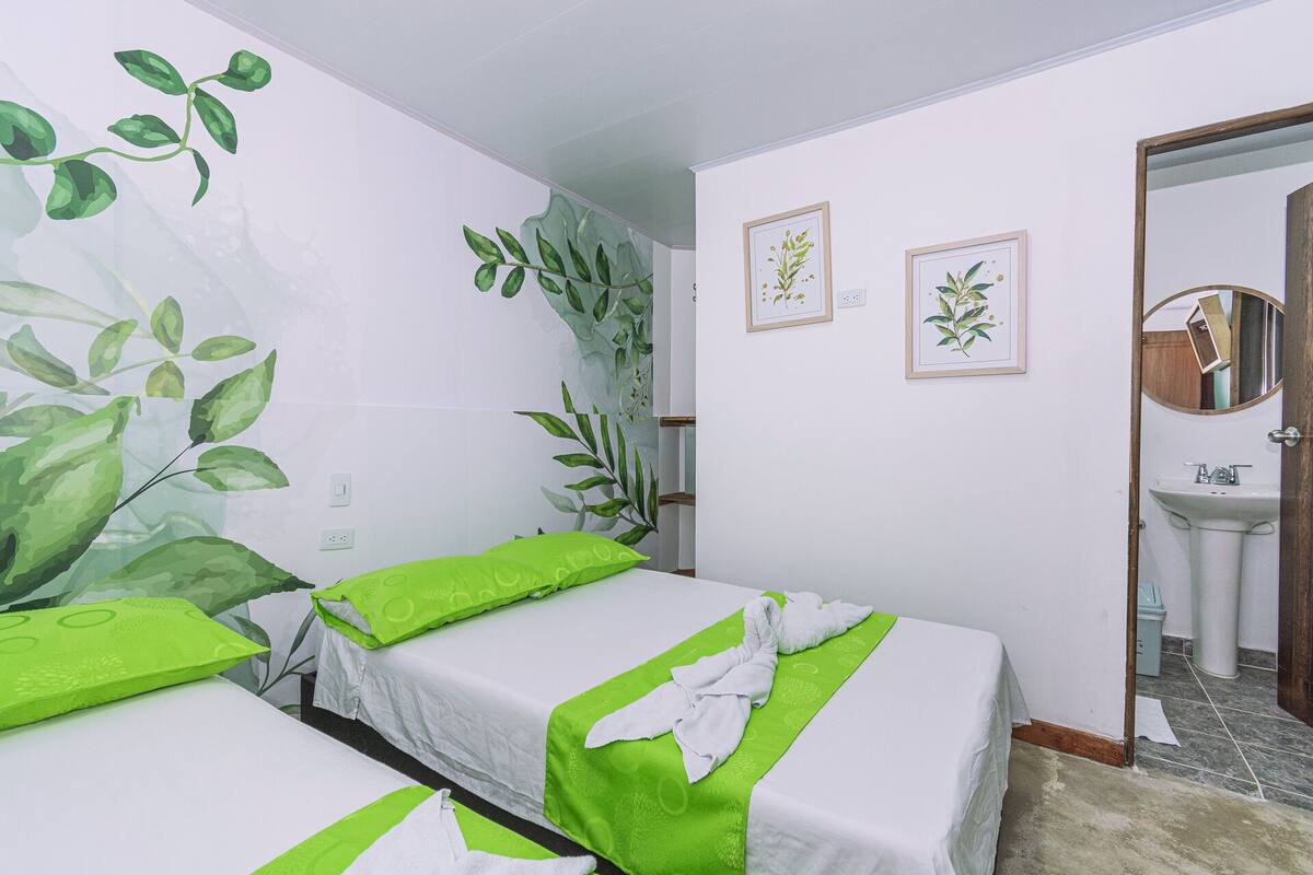 A hotel room with two beds, green pillows, a large wall mural of leaves, and a bathroom with a sink and mirror.