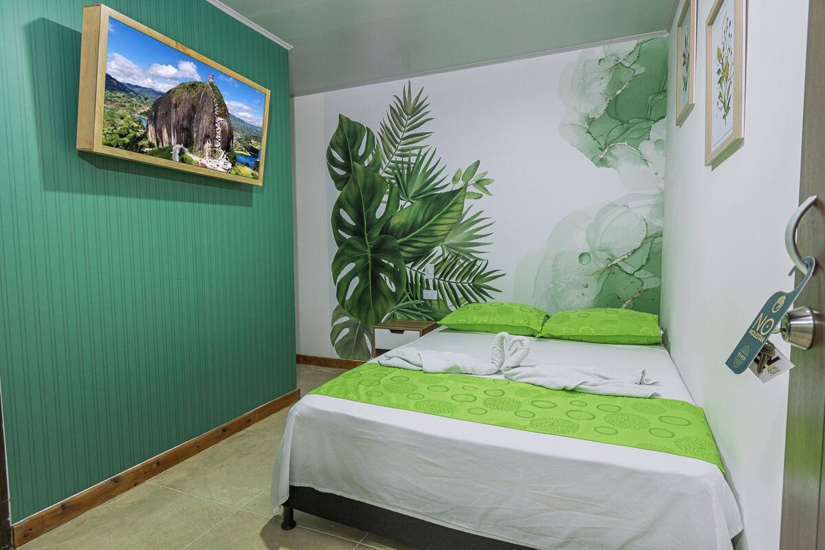 A neatly made bed with a green and white bedspread, a large green plant beside it, and a framed picture of a landscape on the wall.