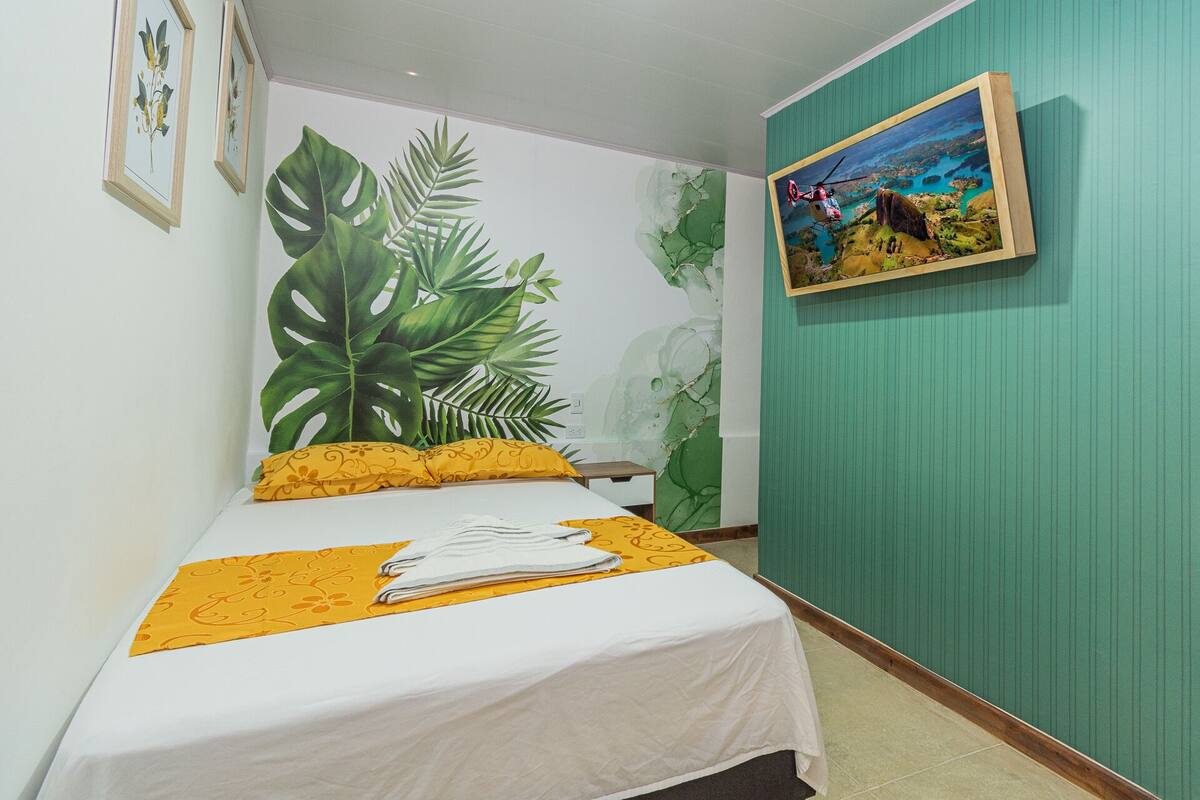 A bedroom with a bed, a large tropical plant mural, and a framed picture on the wall.