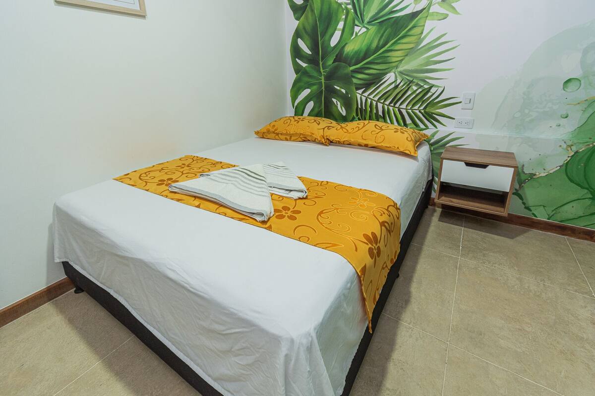 A neatly made bed with a white and yellow bedspread, a folded towel, and a nightstand with a drawer.