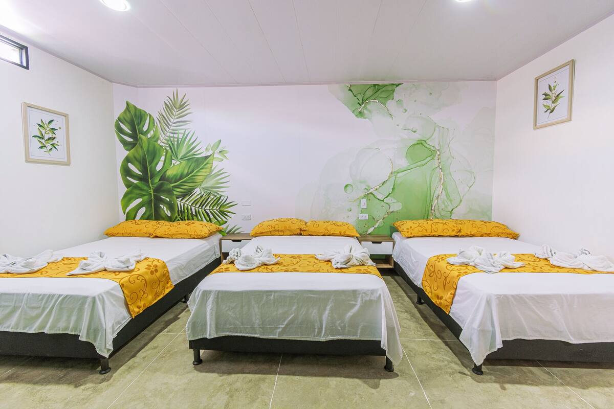 A hotel room with three single beds, each with a white bedspread and yellow pillows. The room features a large wall mural with tropical foliage and a woman.