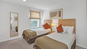 A hotel room with two beds, a mirror, a window with blinds, a lamp, and a framed artwork on the wall.
