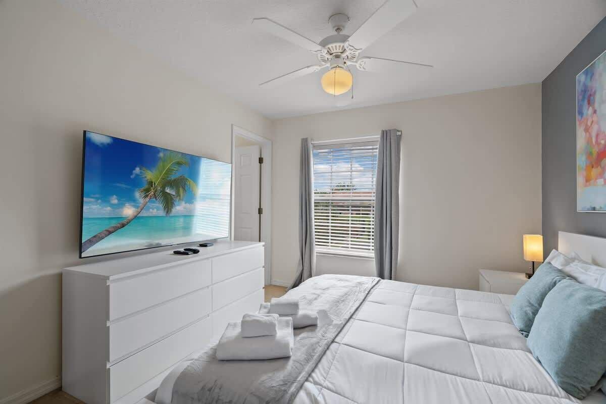 A bedroom with a bed, a TV on the wall, a ceiling fan, and a window with curtains.