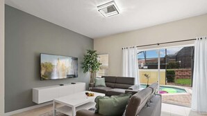 Een moderne woonkamer met een flatscreen-tv, een witte salontafel, een grijze bank en een zwembad zichtbaar door grote glazen deuren.