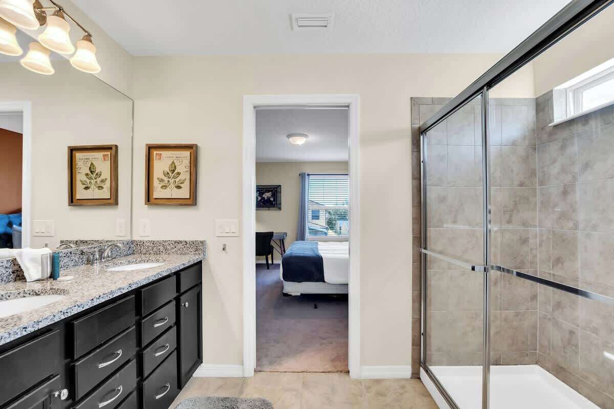 A bathroom with a glass shower enclosure, a sink with a granite countertop, and a mirror with framed artwork.
