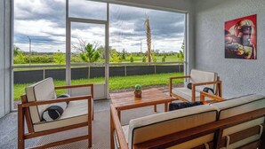 A room with a large window, a wooden table, two chairs, a sofa, and a wall-mounted artwork.