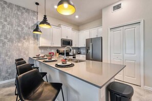 Cozinha moderna com ilha central, armários brancos, eletrodomésticos de aço inoxidável e banquetas pretas.