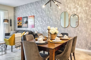 Uma sala de jantar com mesa de madeira posta para quatro pessoas, rodeada por cadeiras. Há espelhos fixados na parede e cartazes emoldurados.