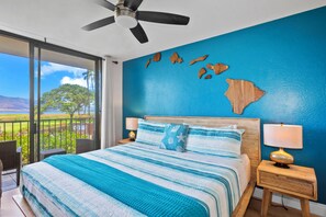 A bedroom with a large bed, a view of the outdoors, a ceiling fan, and a nightstand with a lamp.