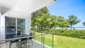 A modern outdoor balcony with a glass door leading to a bedroom, a dining area with a table and chairs, and a view of a beach with palm trees. - Oceanfront Balcony Condo 2101 | Playa Bejuco + Sunset Views + Pool (Bejuco)