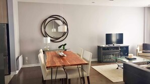 Een moderne eetruimte met een houten tafel en witte stoelen, een ronde spiegel aan de muur, een televisie en een bank in de zithoek.