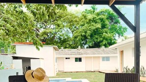 A covered outdoor seating area with wicker furniture, a pool, and a view of a residential building. - Comfy Stay for 2 @CasaSanAlfonso Room #21 (San Alfonso)