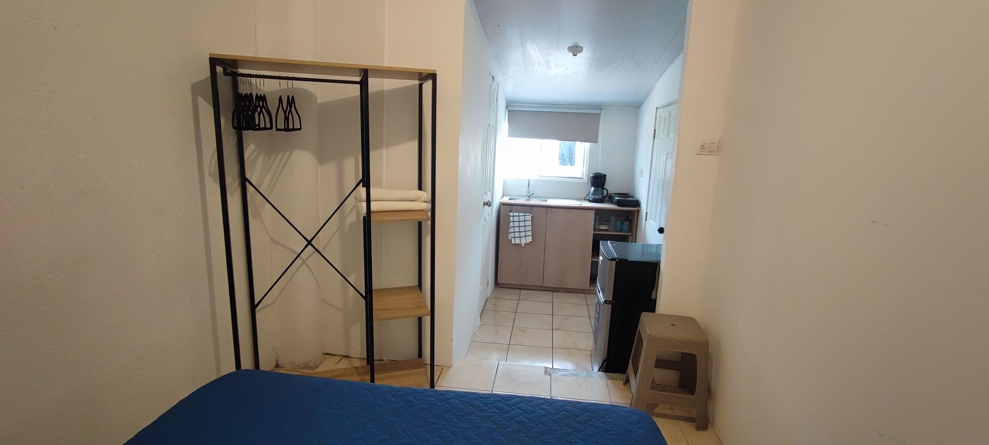 A narrow hallway leading to a bedroom with a bed, a kitchenette with a sink and a stove, and a small stool.