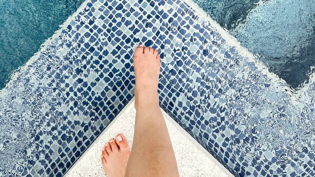 Les pieds d’une personne au bord d’une piscine, avec un motif de mosaïque sur les carreaux.