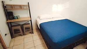 Une petite chambre à coucher, bien rangée, avec un lit simple, une étagère sur laquelle se trouve une machine à café et un tabouret.