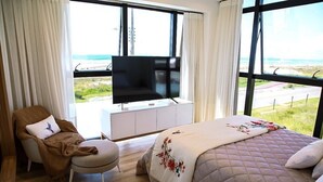 A bedroom with a bed, a TV on a white cabinet, a chair, and a view of the beach. - Apto Pe na Areia - Frente Mar - Imbituba (Imbituba)