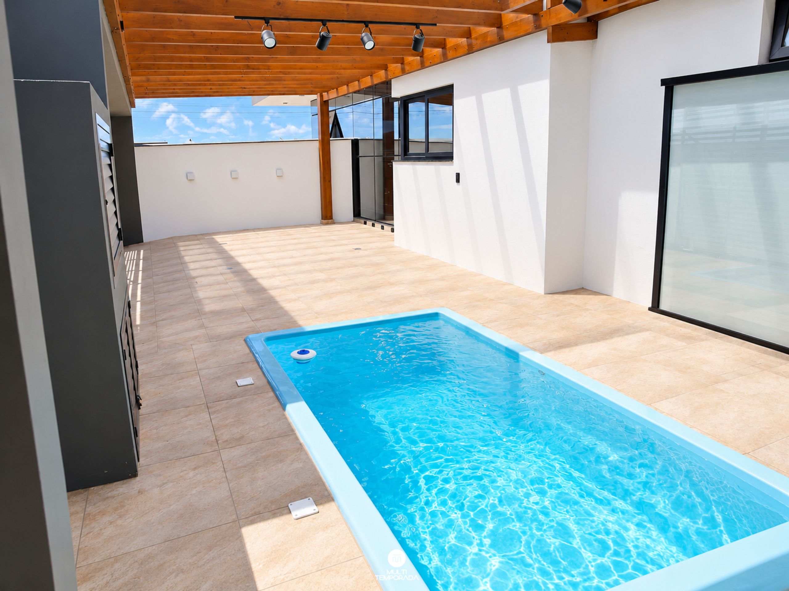 Una piscina exterior moderna con una terraza de madera y una pared de cristal.
