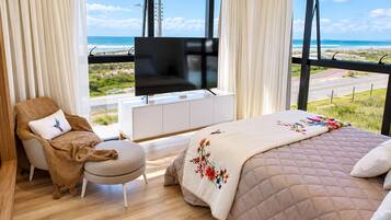 Quarto com cama, TV sobre um armário branco, poltrona com almofada e vista para a praia por grandes janelas.
