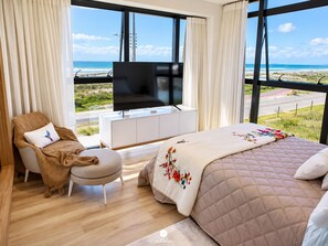Een slaapkamer met een bed, een televisie op een witte kast, een stoel met een kussen en uitzicht op het strand door grote ramen.