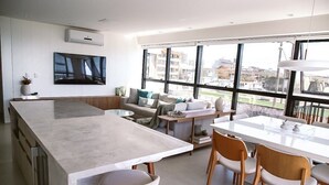 A modern kitchen with a large island, dining area with chairs, a sofa, and a TV mounted on the wall. - Apto Pe na Areia - Frente Mar - Imbituba (Imbituba)