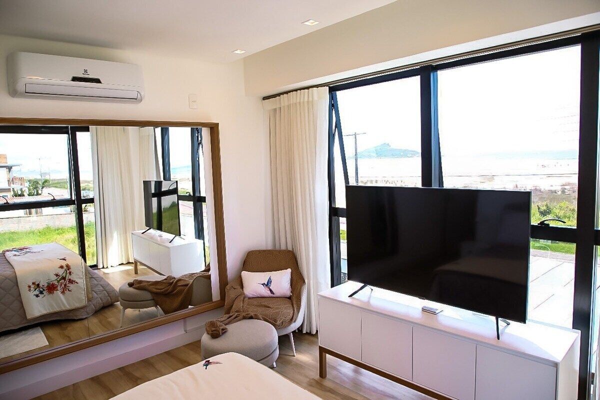 Quarto de hotel com uma janela grande, televisão de tela plana, sofá, poltrona e mesa de cabeceira.
