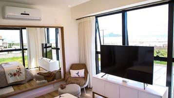 Quarto de hotel com uma janela grande, televisão de tela plana, sofá, poltrona e mesa de cabeceira.