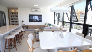 A modern kitchen with a built-in wine rack, a dining area with a large table, and a TV mounted on the wall. - Apto Pe na Areia - Frente Mar - Imbituba (Imbituba)