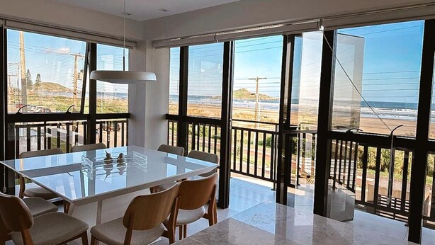 A modern dining area with a glass table and chairs, large windows with a view of the beach, and a balcony with a railing. - Apto Pe na Areia - Frente Mar - Imbituba (Imbituba)