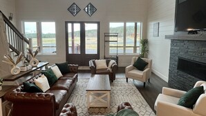 A spacious living room with a high ceiling, a fireplace, and large windows offering views of the outdoors. - “Stay in this brand new luxurious cabin in Hochatown.  (Broken Bow)