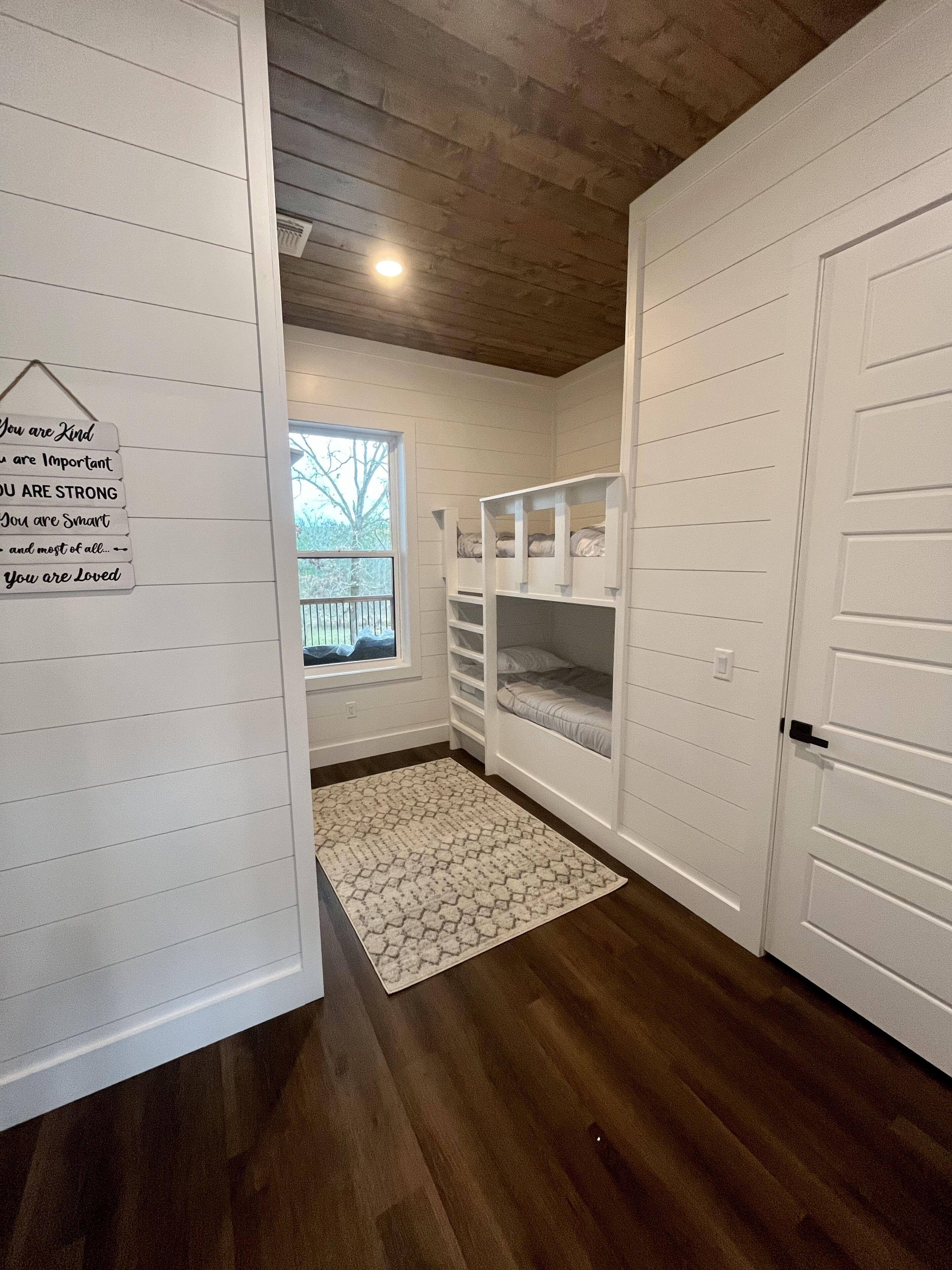 A room with white paneled walls, a wooden ceiling, and a window with a view of trees. There are bunk beds with storage drawers and a white rug on the floor.