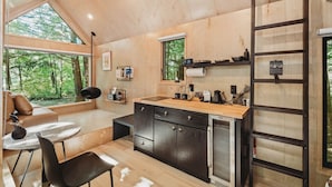 Eine kleine Küche mit einer Holzausstattung, einer integrierten Spüle und einem kleinen Kühlschrank. Ein Fenster bietet Blick ins Freie, und eine Leiter lehnt an der Wand.