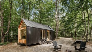 Una cabaña pequeña con techo de metal y paredes de madera, rodeada por un bosque. Hay dos sillas y un pequeño hogar a la vista.