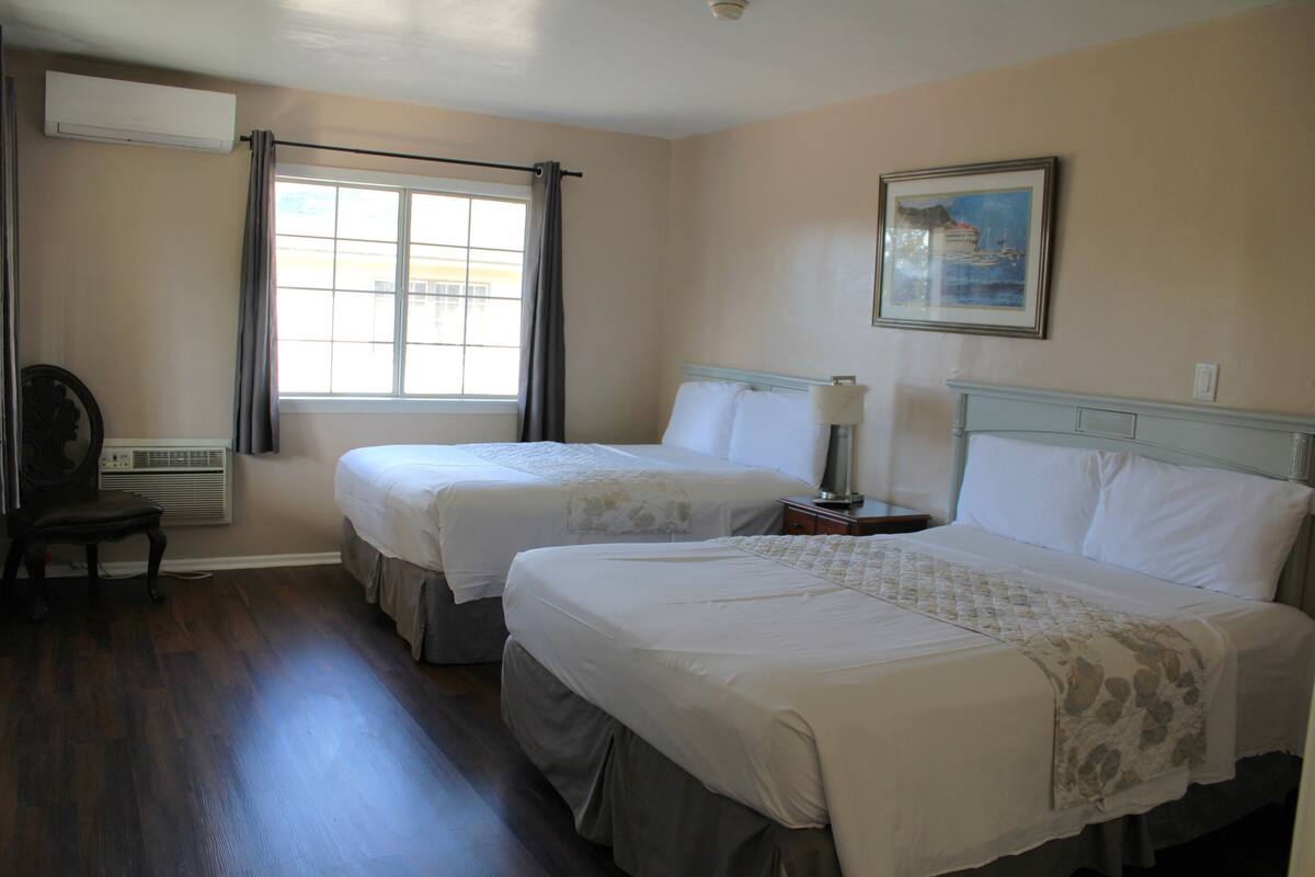 A hotel room with two beds, a window with curtains, a chair, an air conditioner, and a framed picture on the wall.