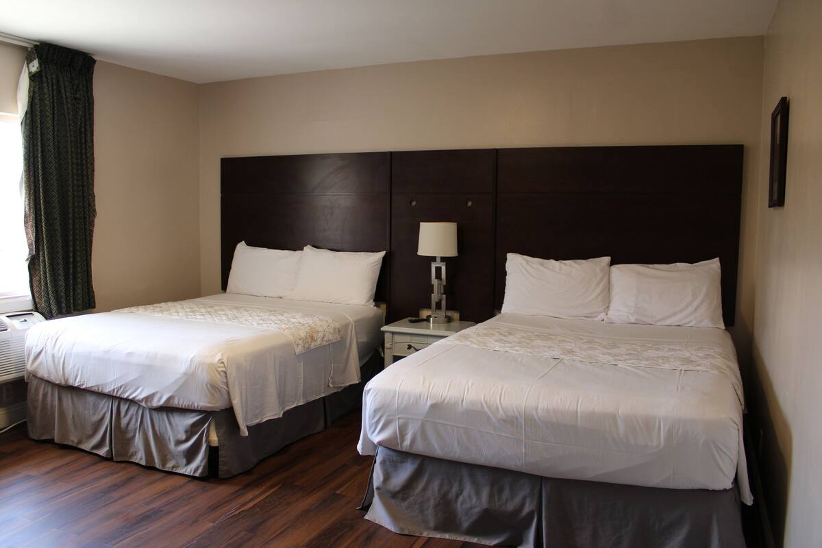 A hotel room with two beds, a nightstand, a lamp, and a window with curtains.