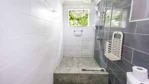 A bathroom with white tiles, a shower, a window with greenery, and a toilet with a paper towel holder.