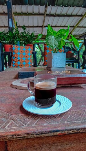 A table with a glass of dark beverage, potted plants, and a patterned cushion. - Rustic and unique private room with large wraparound balcony up in the Trees! (Aburi)