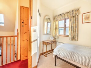 Quarto com porta de madeira, uma mesinha com relógio, cama com roupa de cama listrada e janela com cortinas.