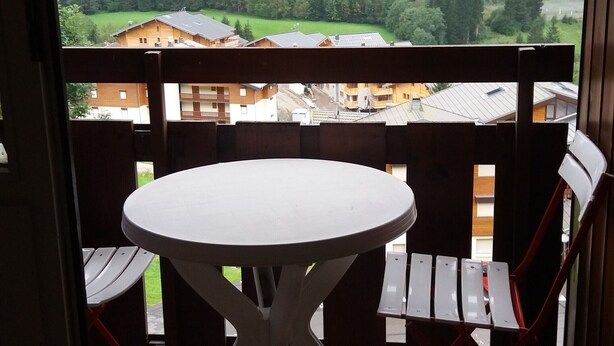 Ein Balkon mit rundem Tisch und zwei Stühlen, der einen Blick auf eine bewaldete Gegend mit Gebäuden im Hintergrund bietet.