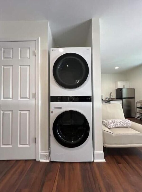 A compact laundry room with two stacked front-loading washing machines, a white door, and a living area with a sofa and a small table.