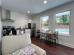 A compact kitchen with modern appliances, a dining area with a wooden table and chairs, and a large window offering a view of the outdoors.