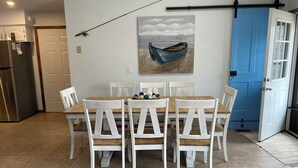 Zona pranzo con tavolo in legno e sedie bianche, un quadro raffigurante una barca sull'acqua, una porta blu e una cucina con frigorifero.