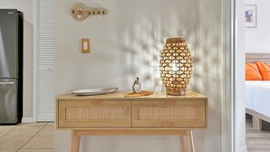 Una habitación con una consola de madera, un letrero de bienvenida con forma de llave decorativa, una lámpara de fibras naturales y una cama con almohadones naranjas.